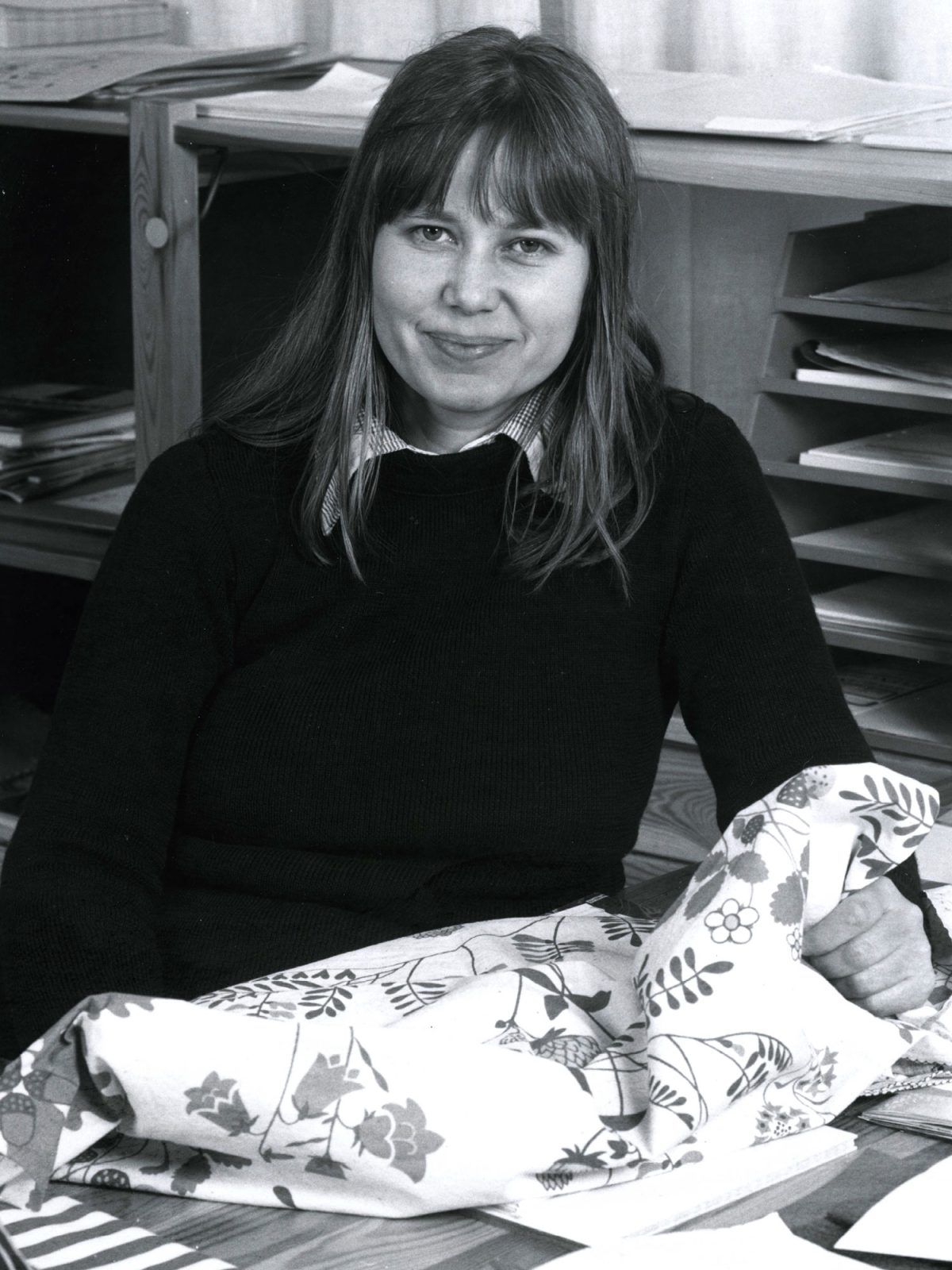 Ung leende kvinna med långt hår håller i tyg med mönster av blommor och blad.