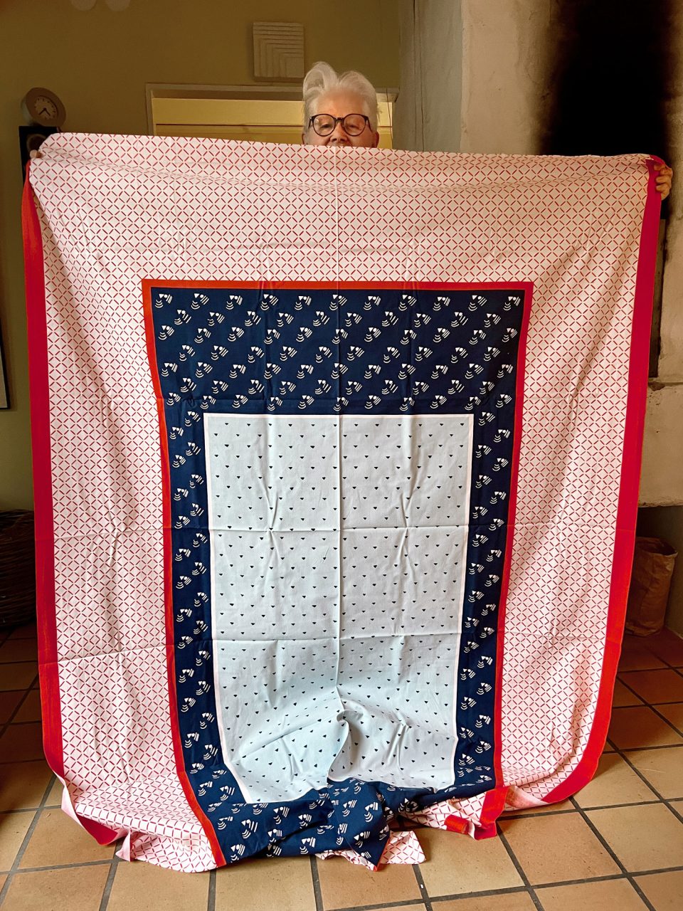 Vithårig kvinna, Vivianne Sjölin, håller lapptäcke framför sig, gjort av olika tyger i mönster i blått, rött och vitt.