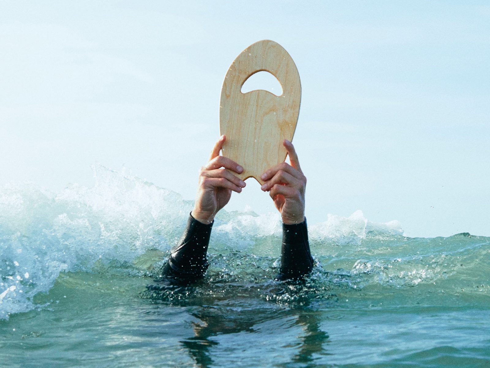 Armar i våtdräkt håller liten träbräda i ljust trä ovanför vattenytan, från IKEA KÅSEBERGA surfkollektion.