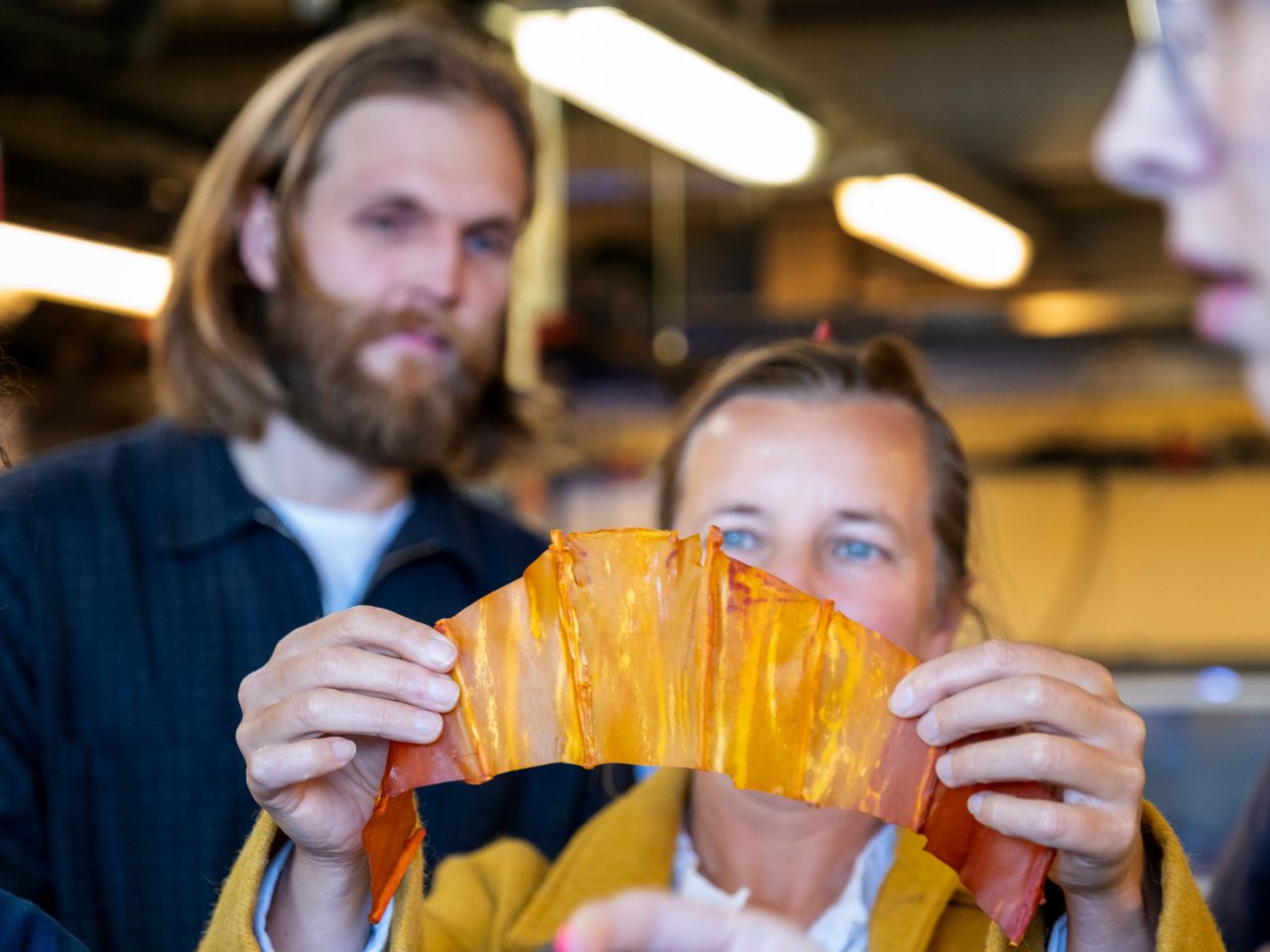 Kvinna håller upp en liten bit glansigt, guldfärgat tunt material, en skäggig man tittar på