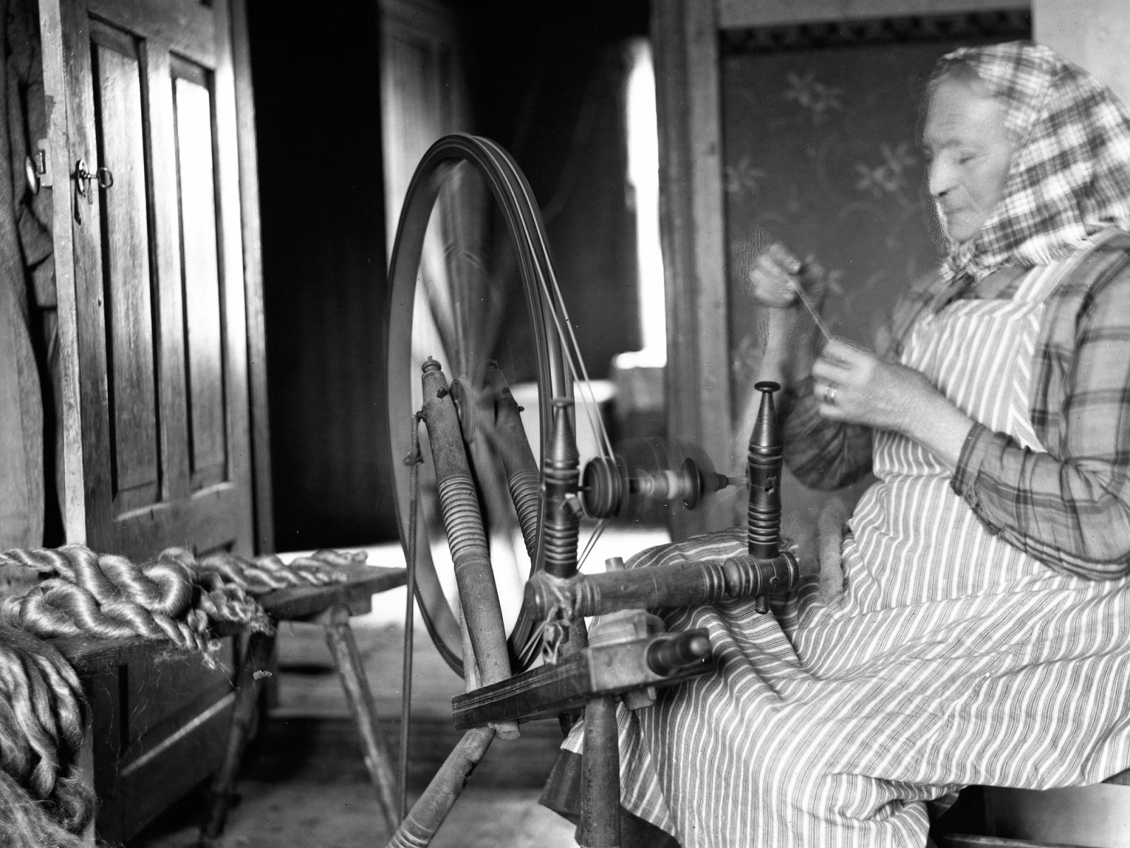 Blond kvinna i sjal och randigt förkläde spinner lin på en spinnrock, 1920-tal.