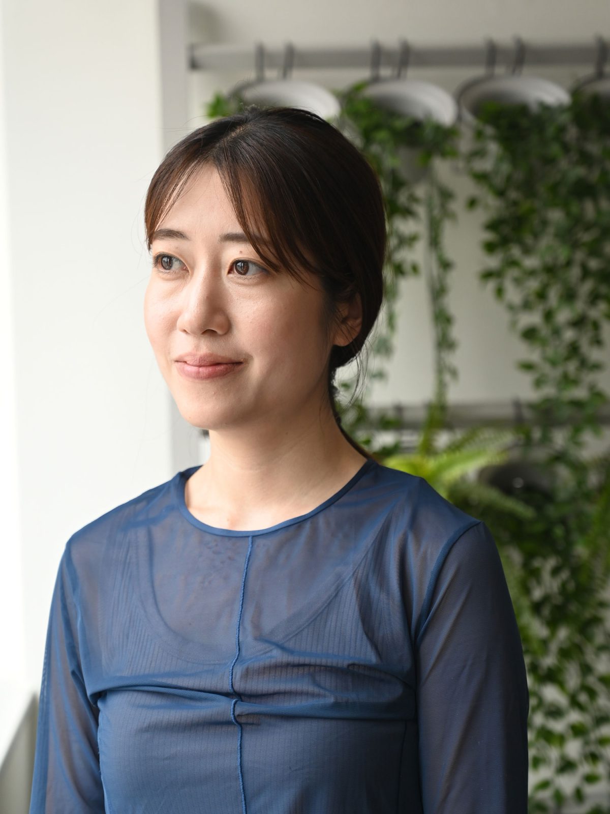 Mörkhårig kvinna i blå blus, Mai Tanigawa, står framför en vägg med gröna växter.