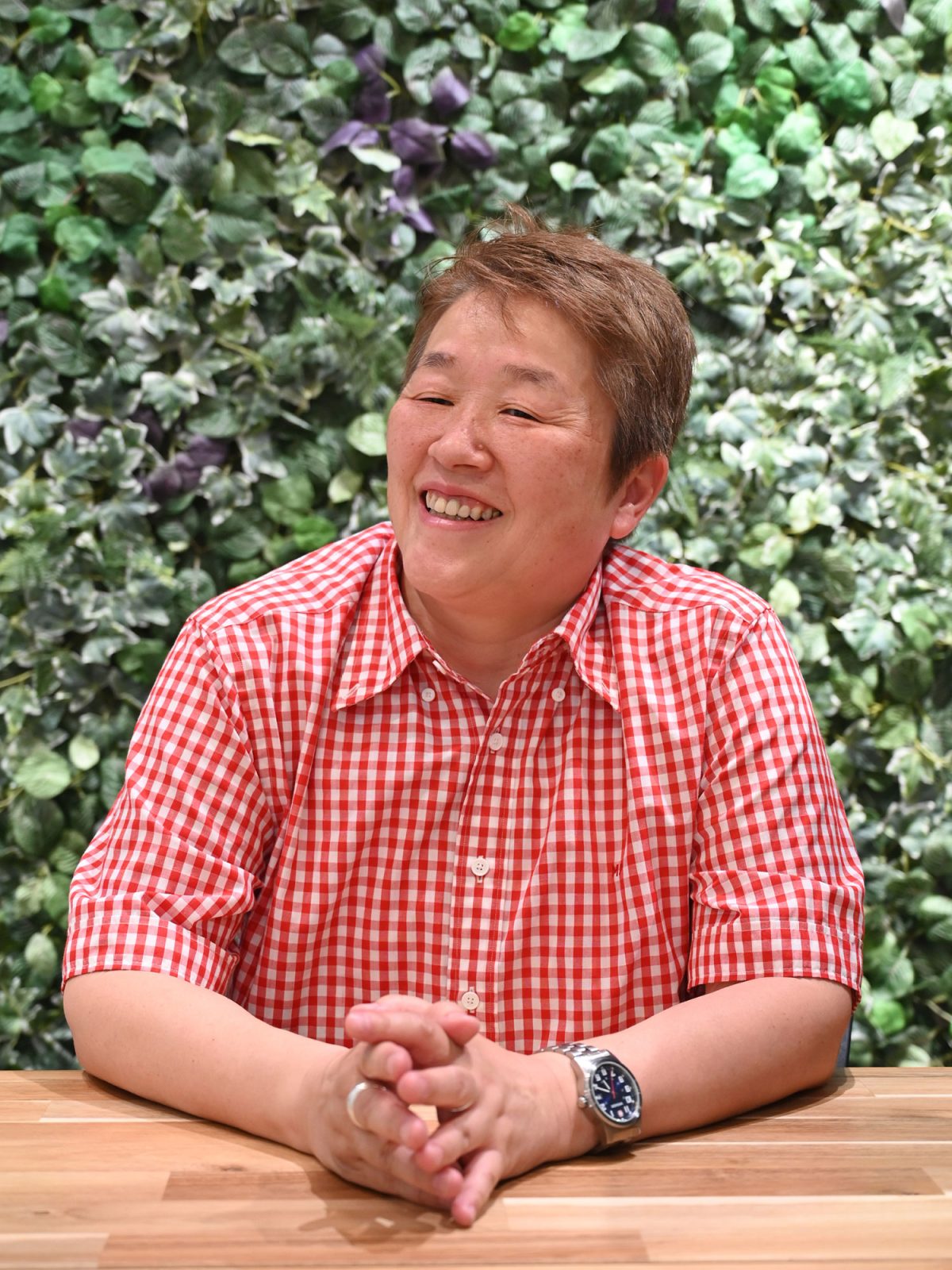 Leende mörkblond kvinna, Tamae Asayama, i röd-vitrutig skjorta sitter framför en vägg med gröna växter.