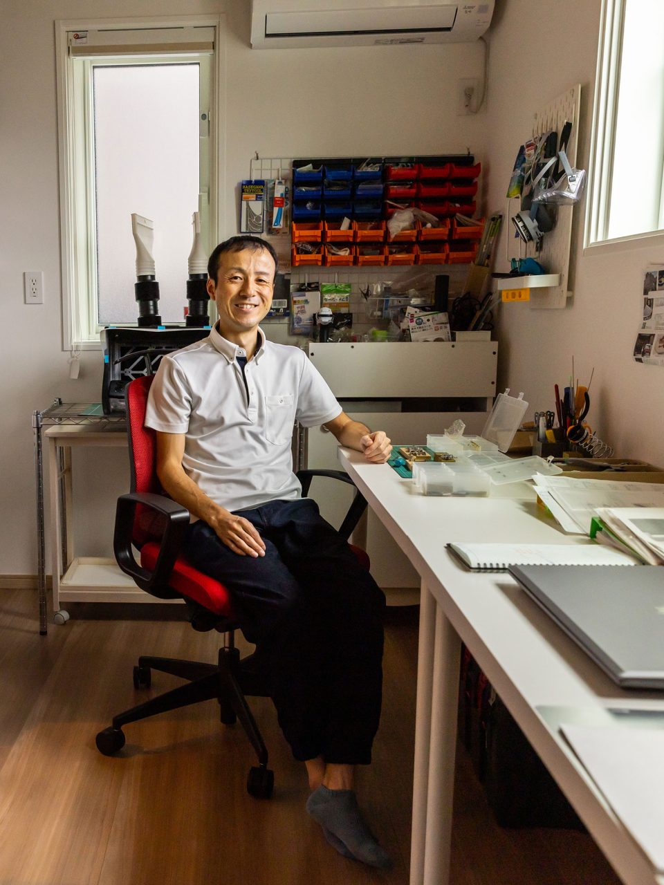 小さな工具やネジがしまわれているウォールシェルフの前で、白い長テーブルの横に座る黒髪の男性。