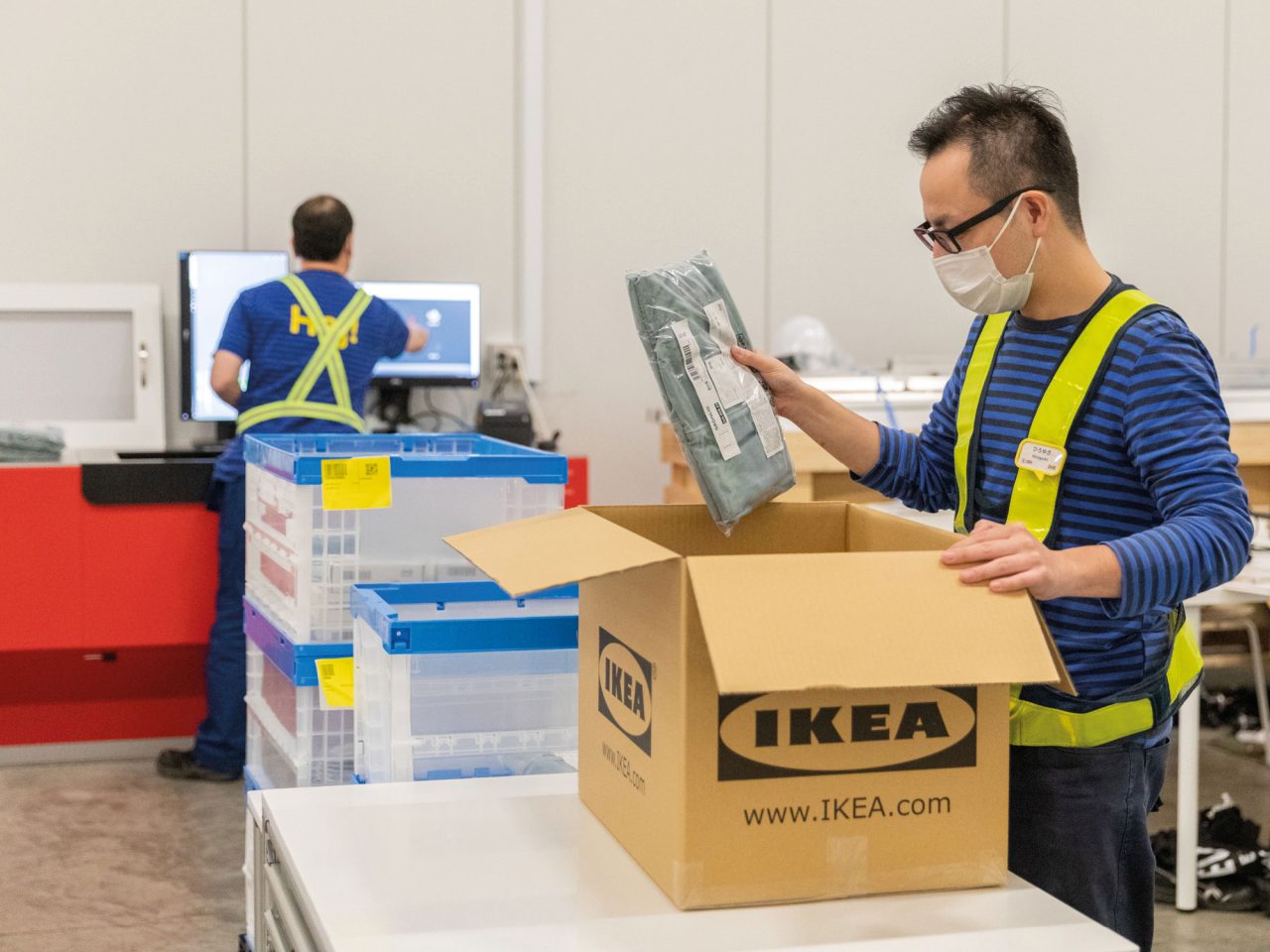 黒髪のイケアのコワーカーが茶色のダンボールに小物を詰め、別のコワーカーはボックスとコンピューターのスクリーンをチェックする。