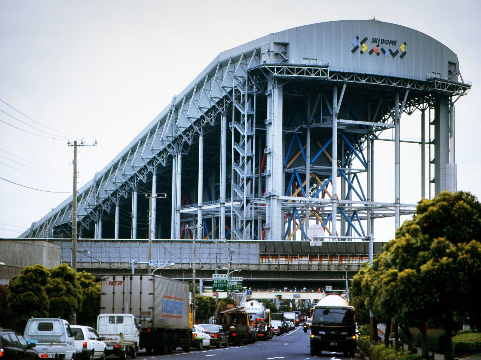 Enorm stålkonstruktion med texten Ski Dome på sidan, en trafikerad motorväg syns i förgrunden.