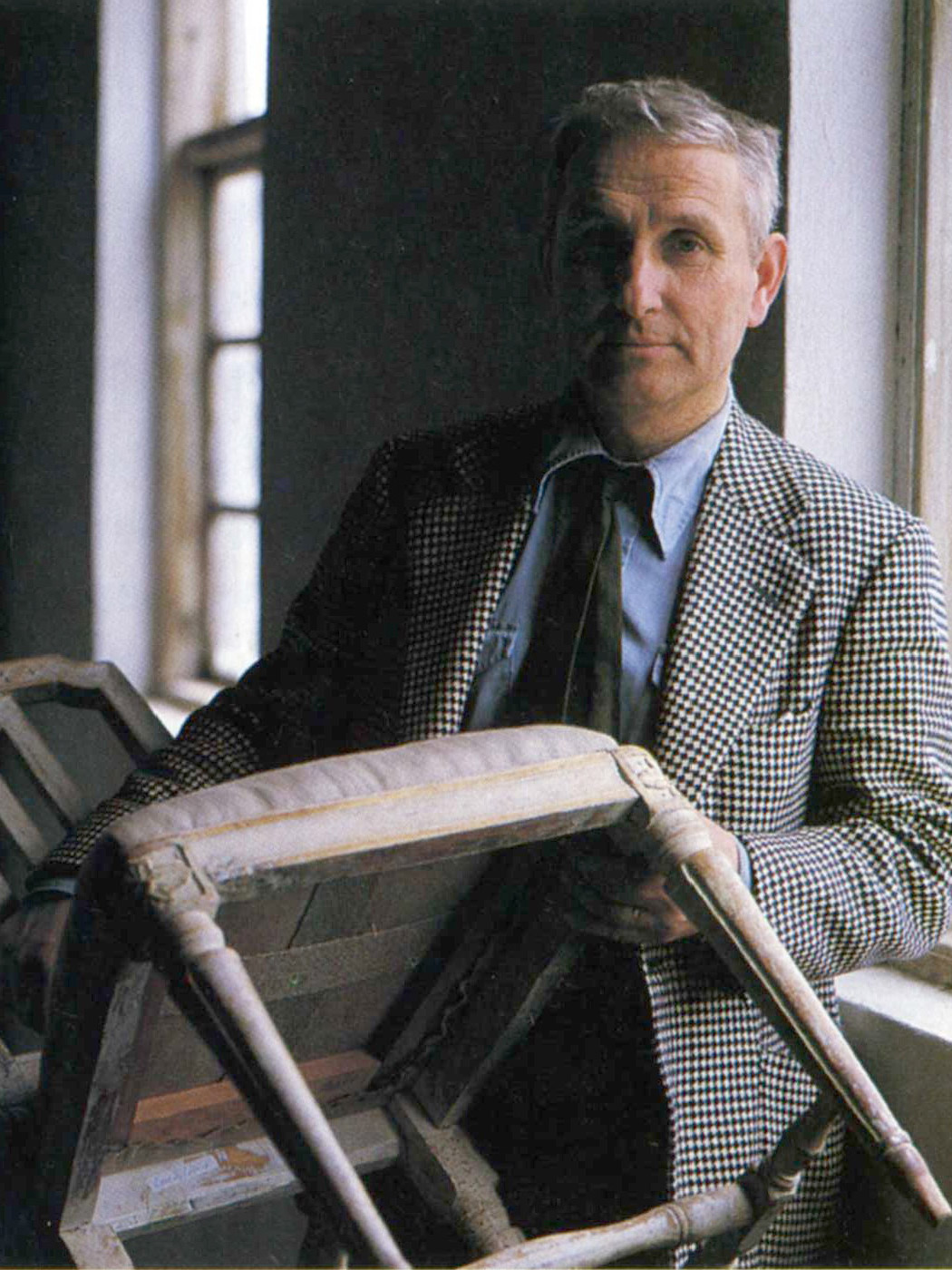 Grey-haired man in check blazer, blue shirt and tie, holding up an older chair.