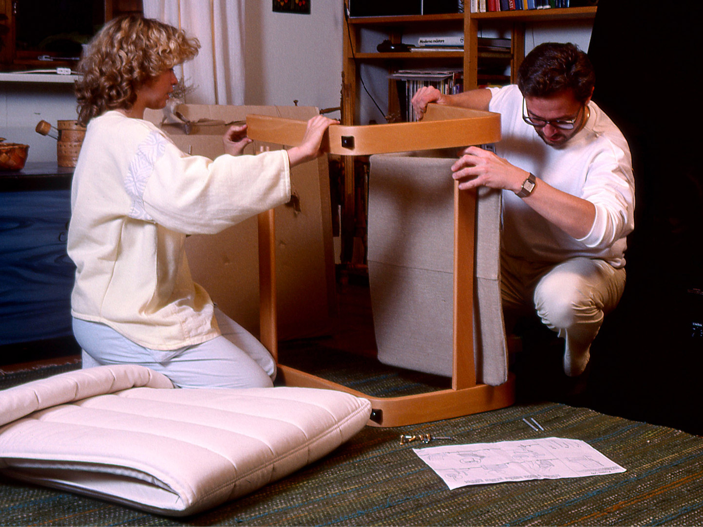 Young couple in 1980s clothes and hairstyles helping each other assemble IKEA furniture in their living room.