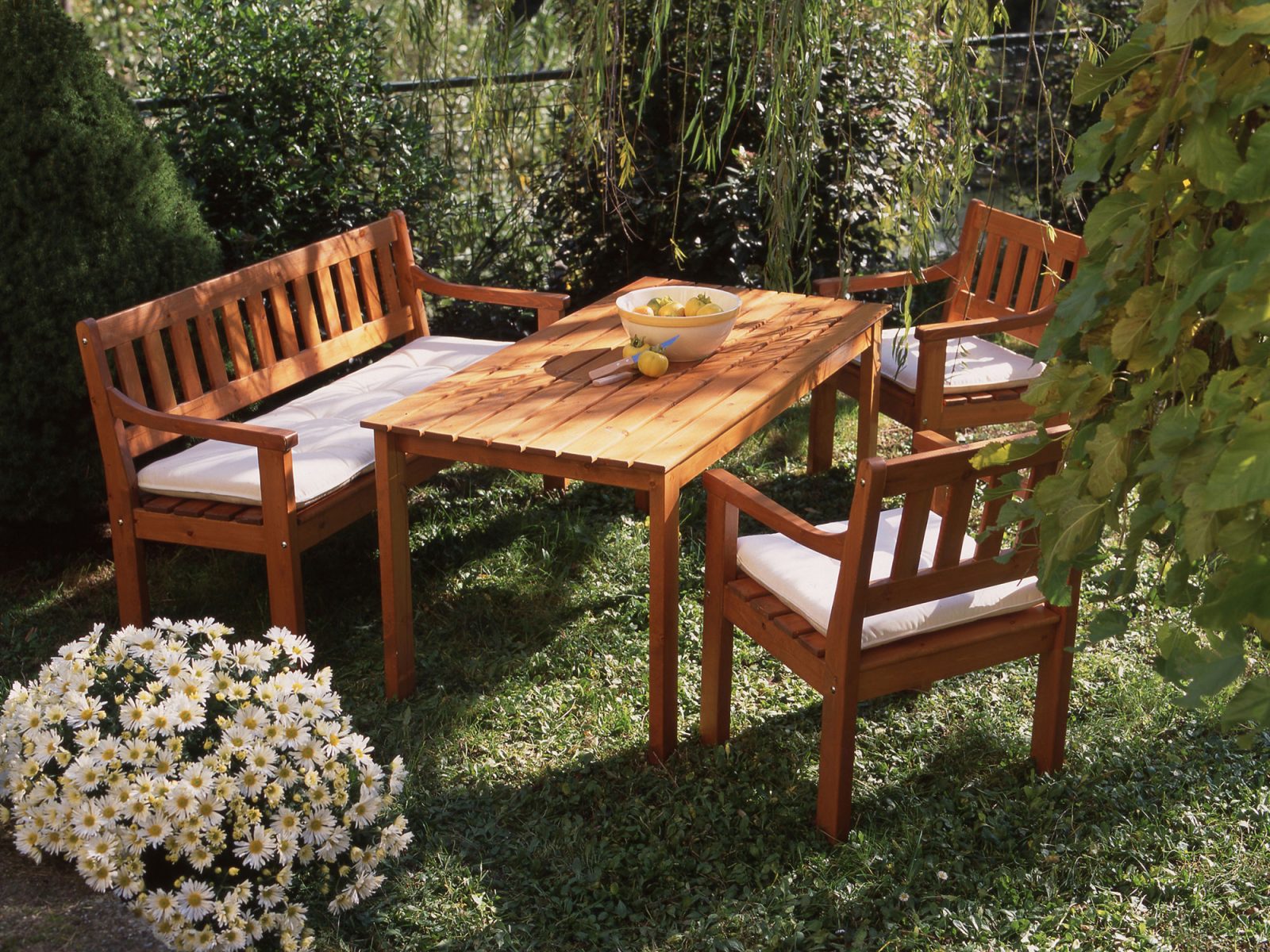 Outdoor furniture in brown wood, SOLHAGA table, sofa, and chairs, are set in a lush garden.