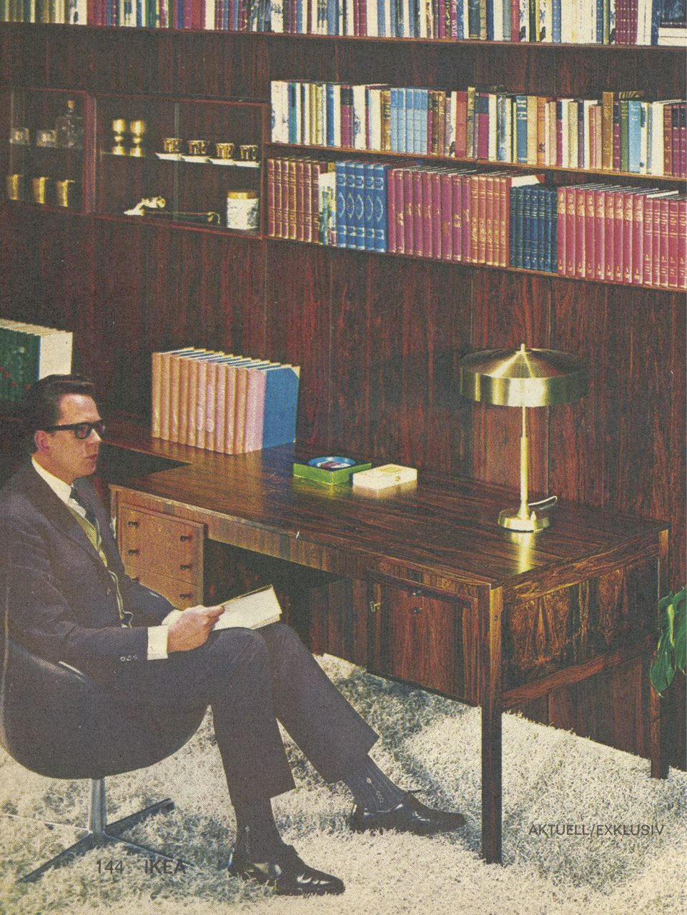 Man in a black suit, tie, white shirt sits in a leather swivel chair before a desk made of rosewood, bookshelf in background.