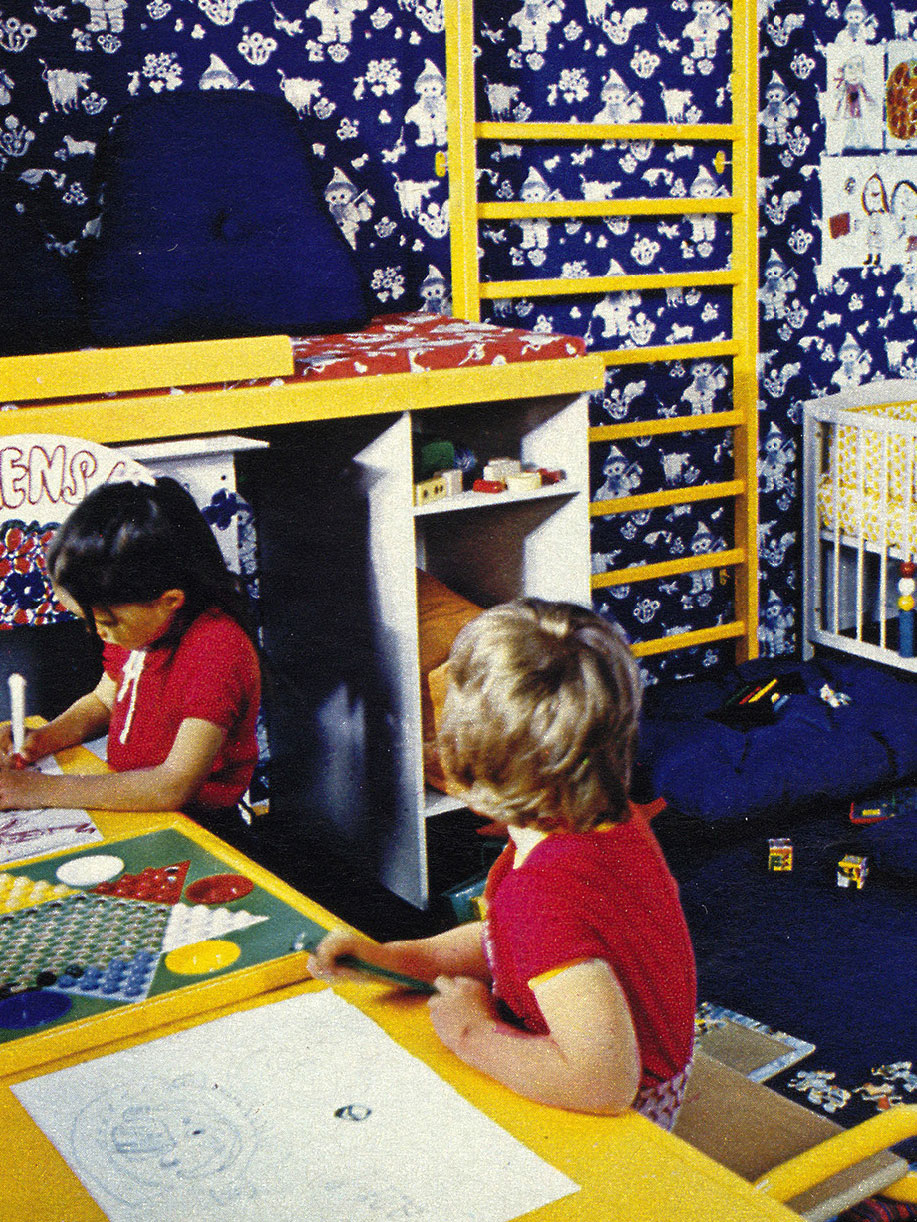 Two children in 1970s clothing sit at a desk drawing and painting, with a wall-mounted gym ladder in the background.