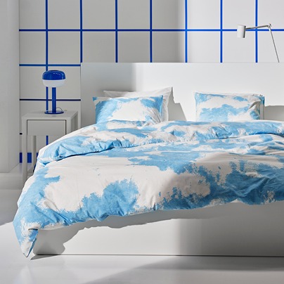 White bed with duvet cover and pillowcases patterned with white clouds on blue sky. Blue lamp on bedside table.
