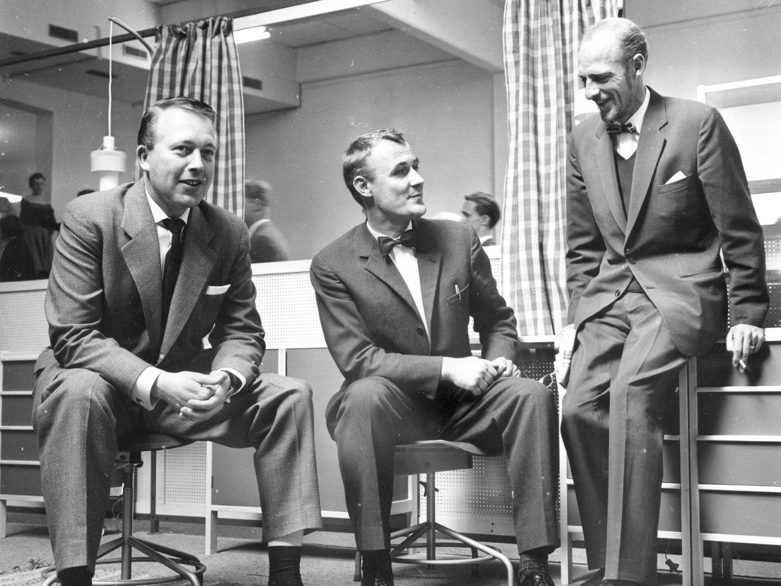 Three smiling men in 1960s suits sit and stand chatting in a furniture store.