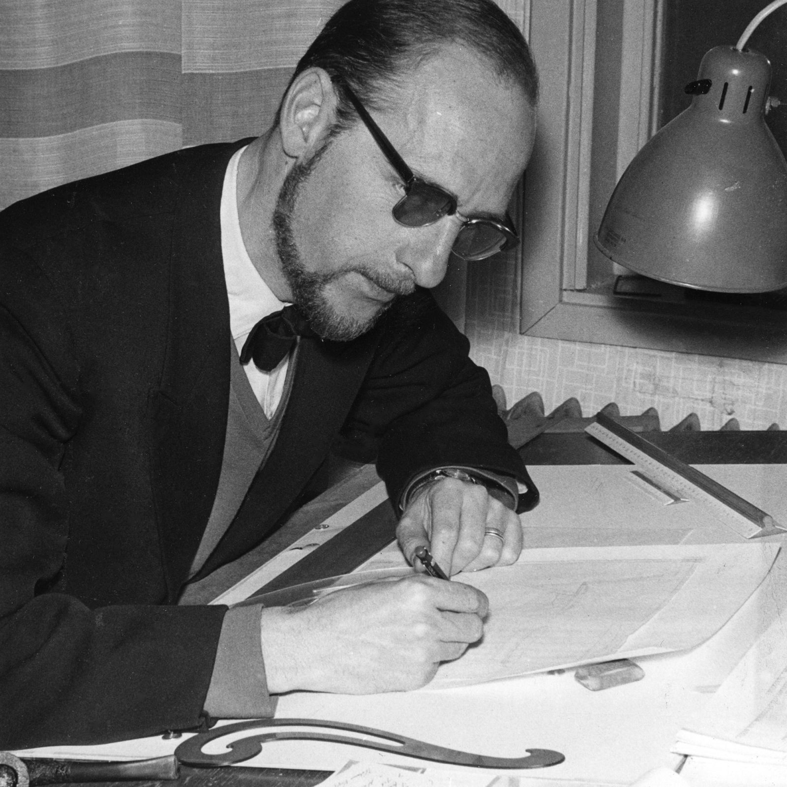 Man in dark glasses and suit sits at a desk, sketching.