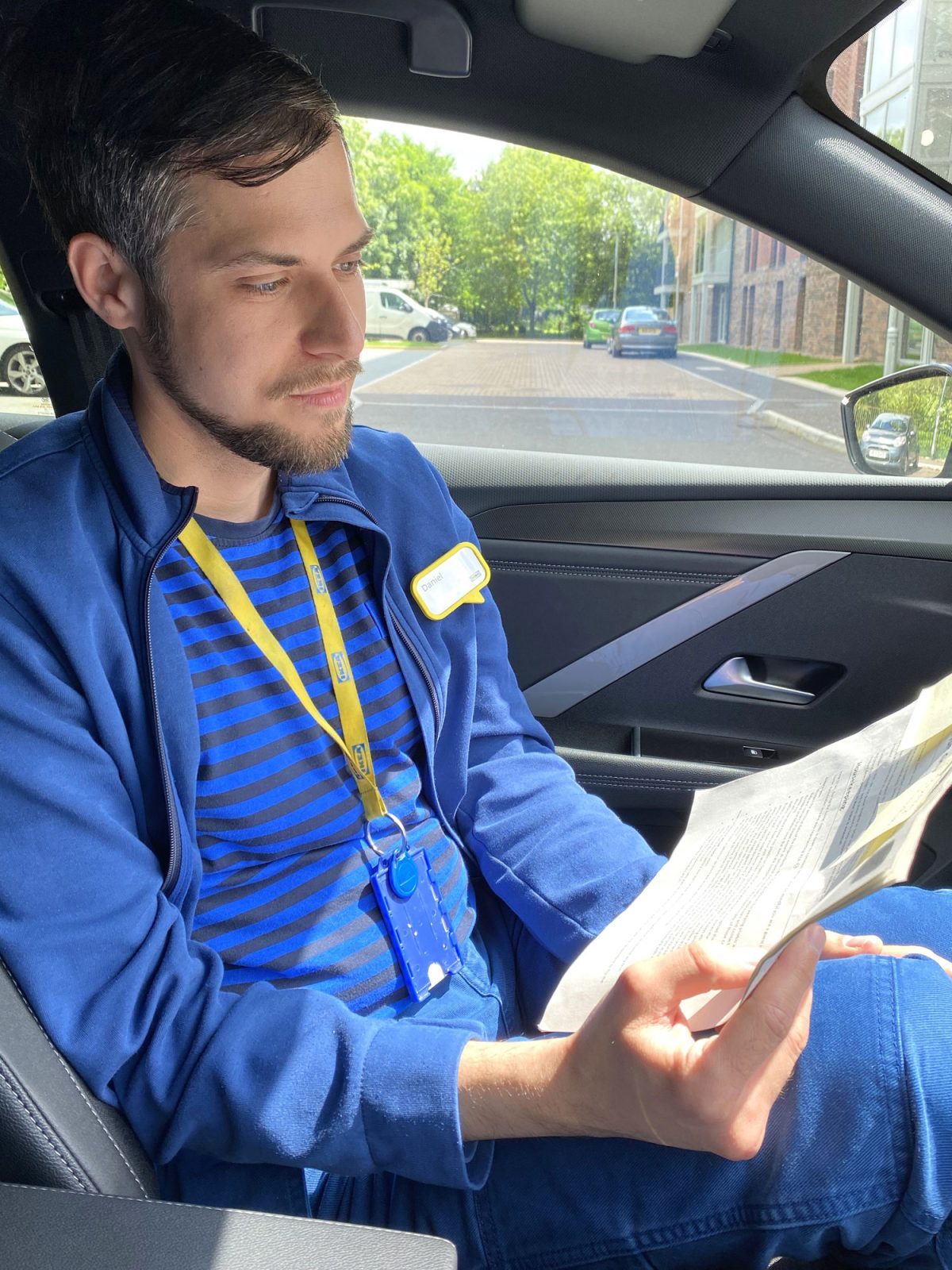 Ung mörkhårig man, Daniel Farago iklädd IKEA arbetskläder sitter i en bil och studerar några papper.
