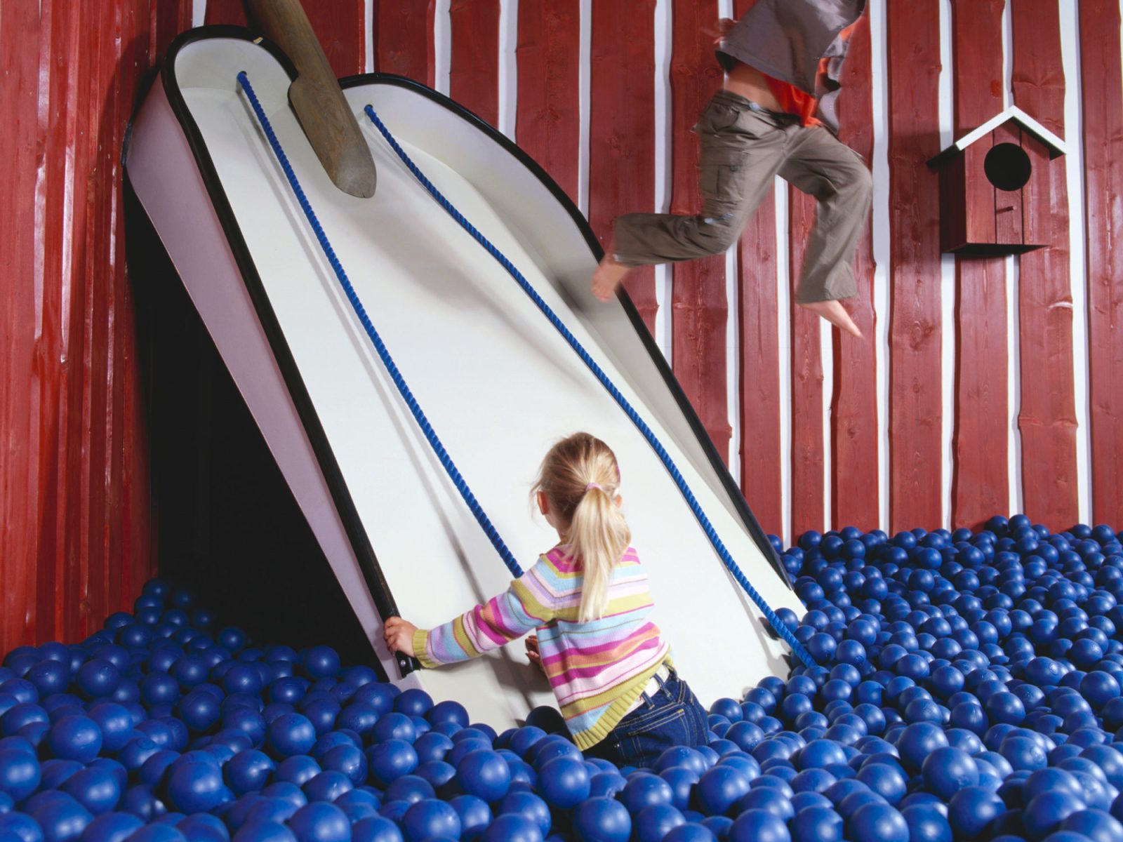 Children playing on a white slide ending in a large ball pit with blue balls.
