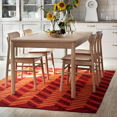 Blond wood dining table and chairs stands on flatwoven orange and red rug with a zigzag pattern.