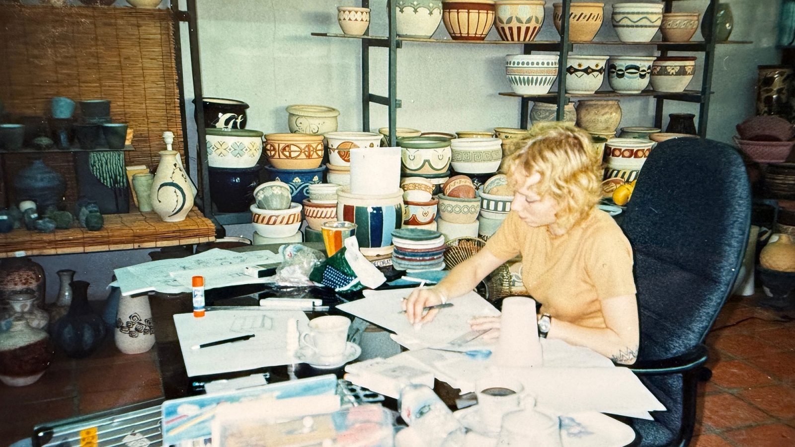 Young blonde woman sits at a desk sketching, clay pots visible in the background.