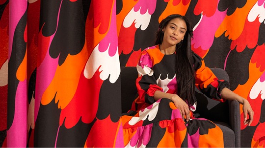 Mörkhårig ung kvinna i klänning med färgglatt mönster i orange, vitt, rosa, rött och svart.