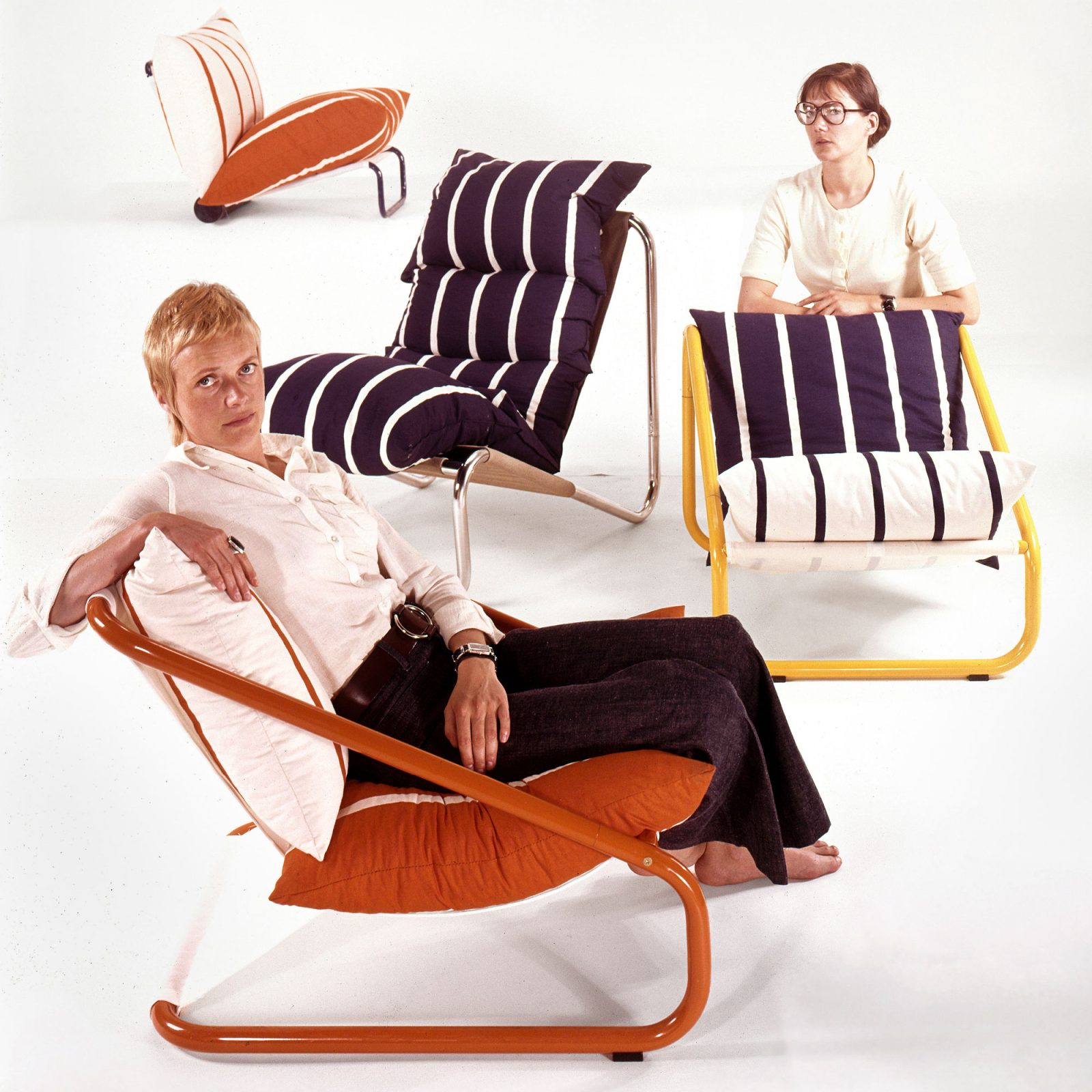Two young women sit in and behind four tube armchairs in different colours and styles, all with striped pillows.