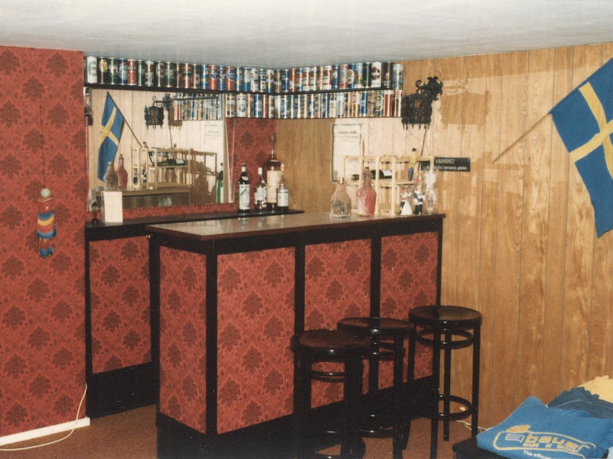 Hembyggd bar med flaskor och glas, en svensk flagga hänger på väggen.