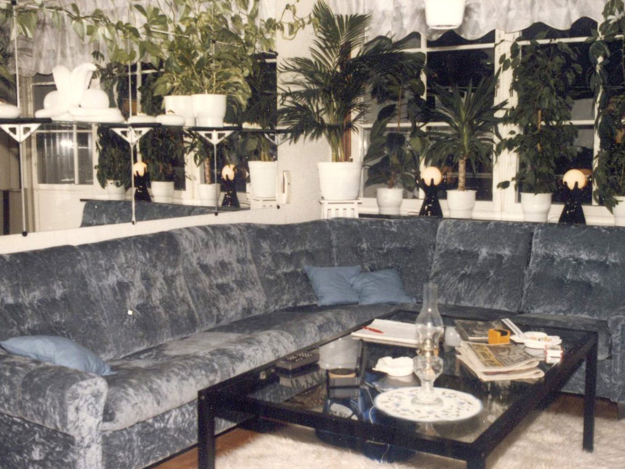 Vardagsrum med stor grå hörnsoffa i sammet under spegelvägg och många gröna växter.