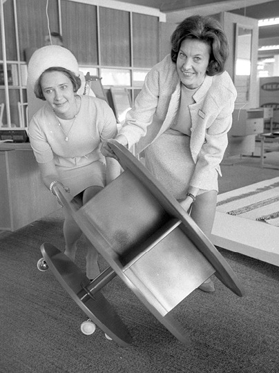 Two women in 1960s clothing carry a round table together, laughing.