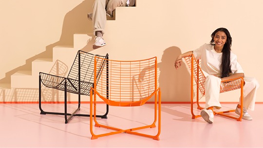 Mörkhårig kvinna sitter i en av tre stolar av stålnät, i orange och svart.