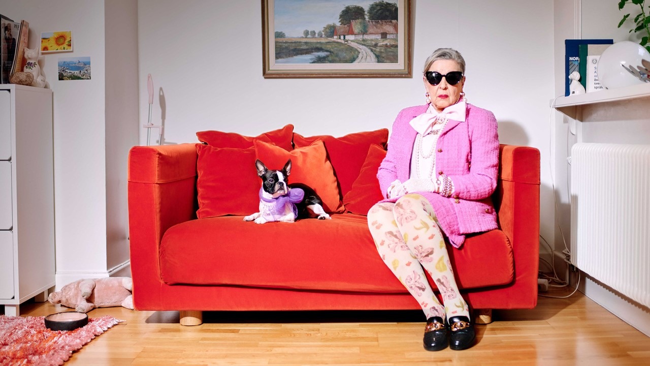 En äldre kvinna med solglasögon, rosa jacka och blommiga strumpbyxor sitter i en orange soffa. Bredvid henne ligger en hund med en lila rosett runt halsen. Ovanför dem hänger en tavla.