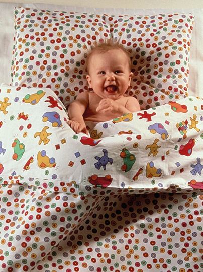 Smiling baby lies in a bed in which the duvet cover has different patterns on each side, LEKLIND.