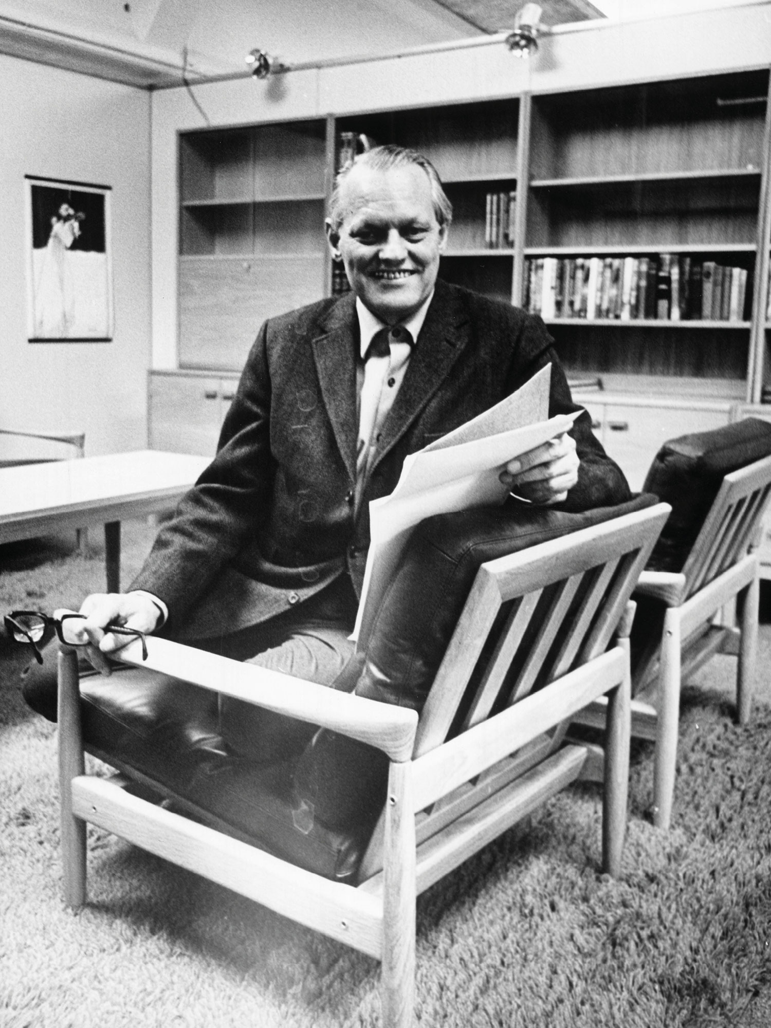 Smiling man dressed in a suit, white shirt and tie sits in an armchair, holding papers, Erik Wørts.