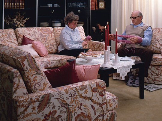 An elderly couple sits in a comfy flowery DJURSHOLM sofa, the man helping the woman with her knitting.