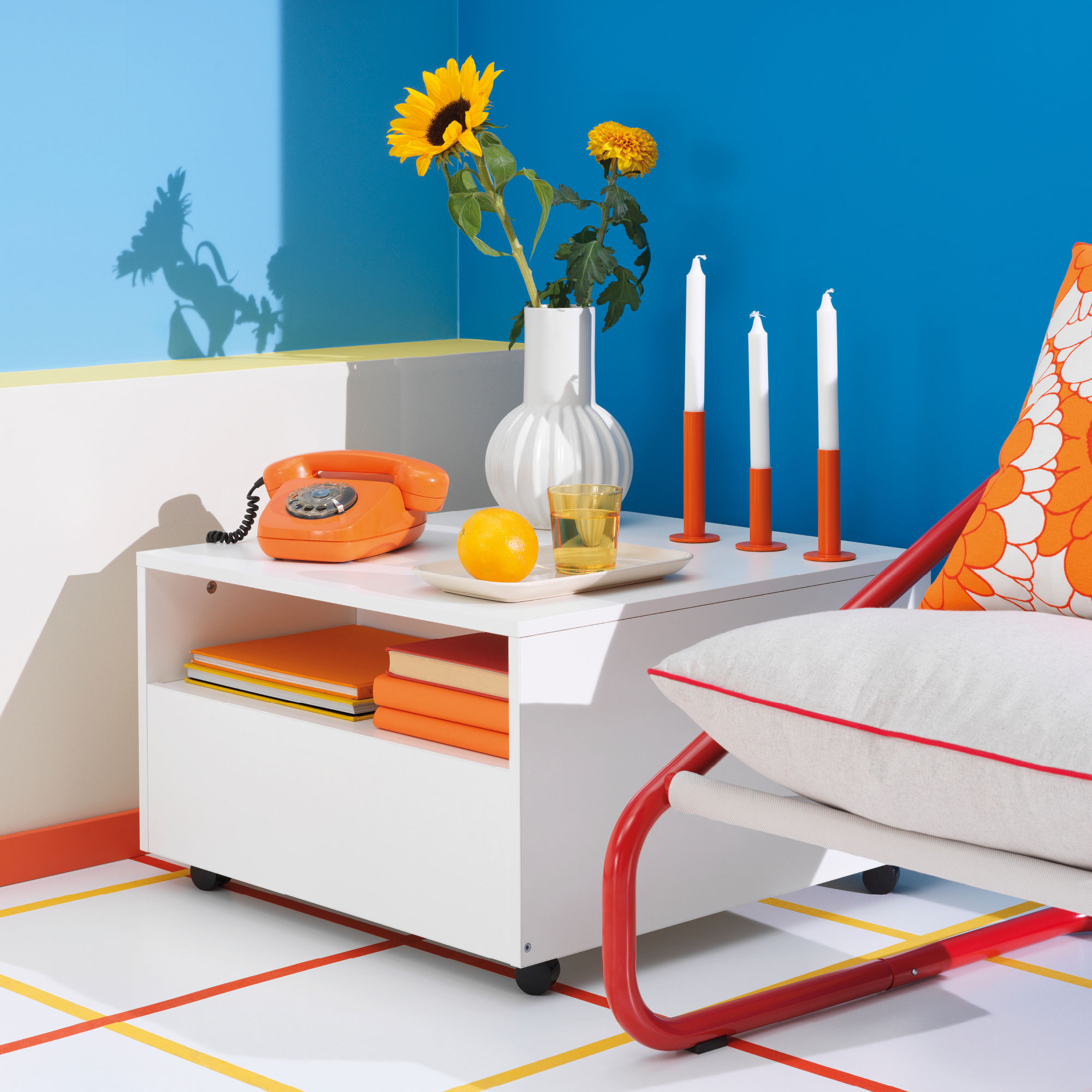 An orange 1960s telephone on a white square coffee table stands next to a low white and red armchair with a steel tube frame.