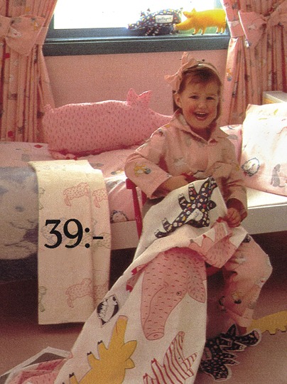 Laughing girl in pink pajamas sits on her bed holding a fabric decorated with drawings of a sow and her piglets.