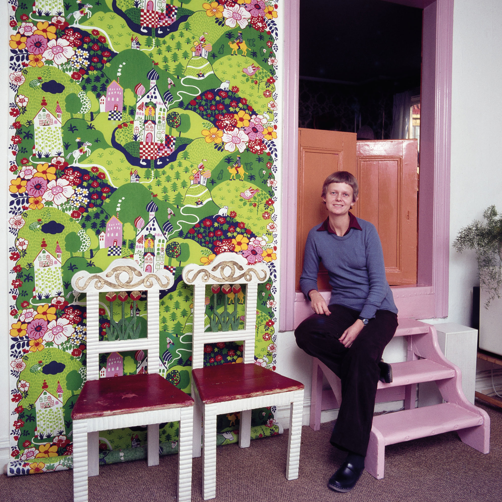 A young woman with a pixie cut sits next to two chairs and a hanging fabric with a colourful, imaginative pattern.